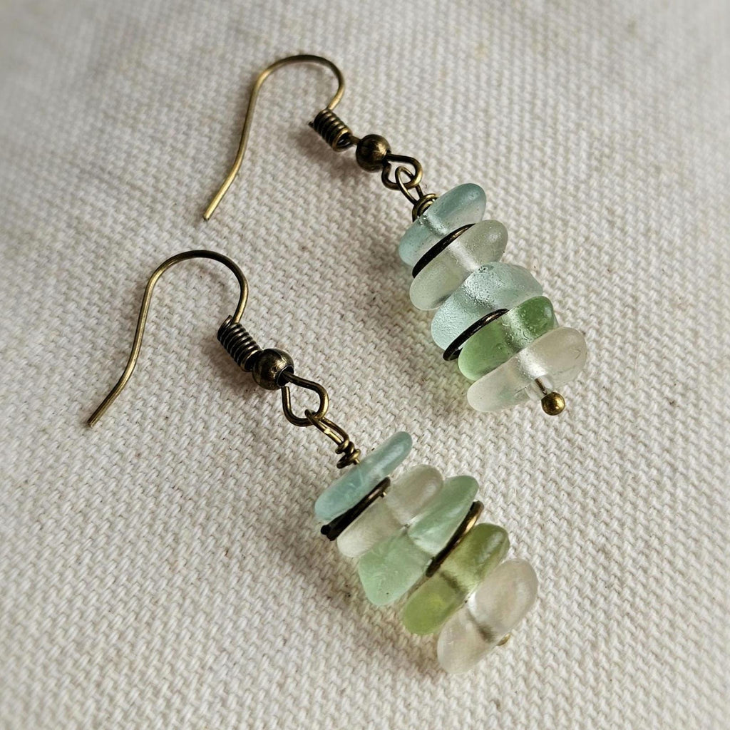 Pair of earrings with green, aqua and clear sea glass on a textured surface