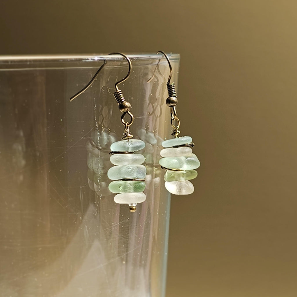 Pair of earrings with green, aqua and clear sea glass on a textured surface dangling from a glass