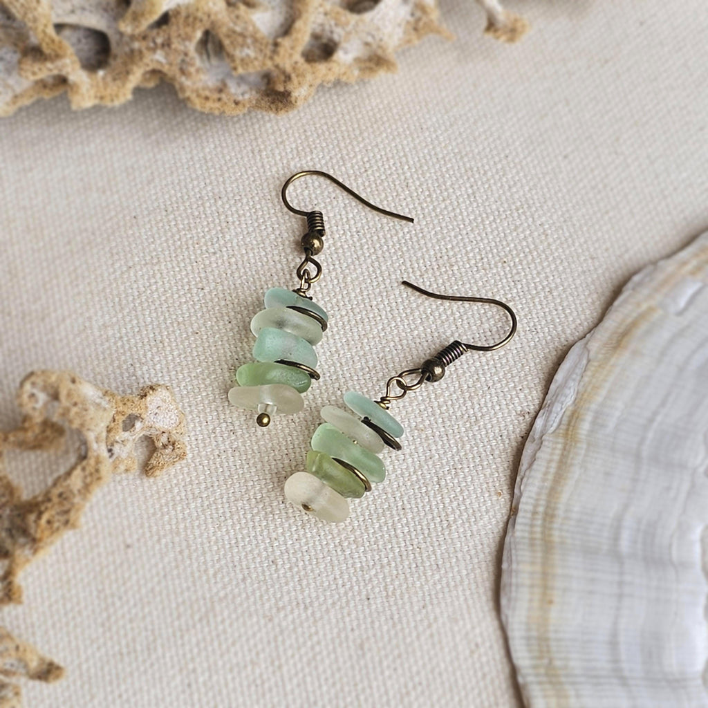 Pair of earrings with green, aqua and clear sea glass on a textured surface