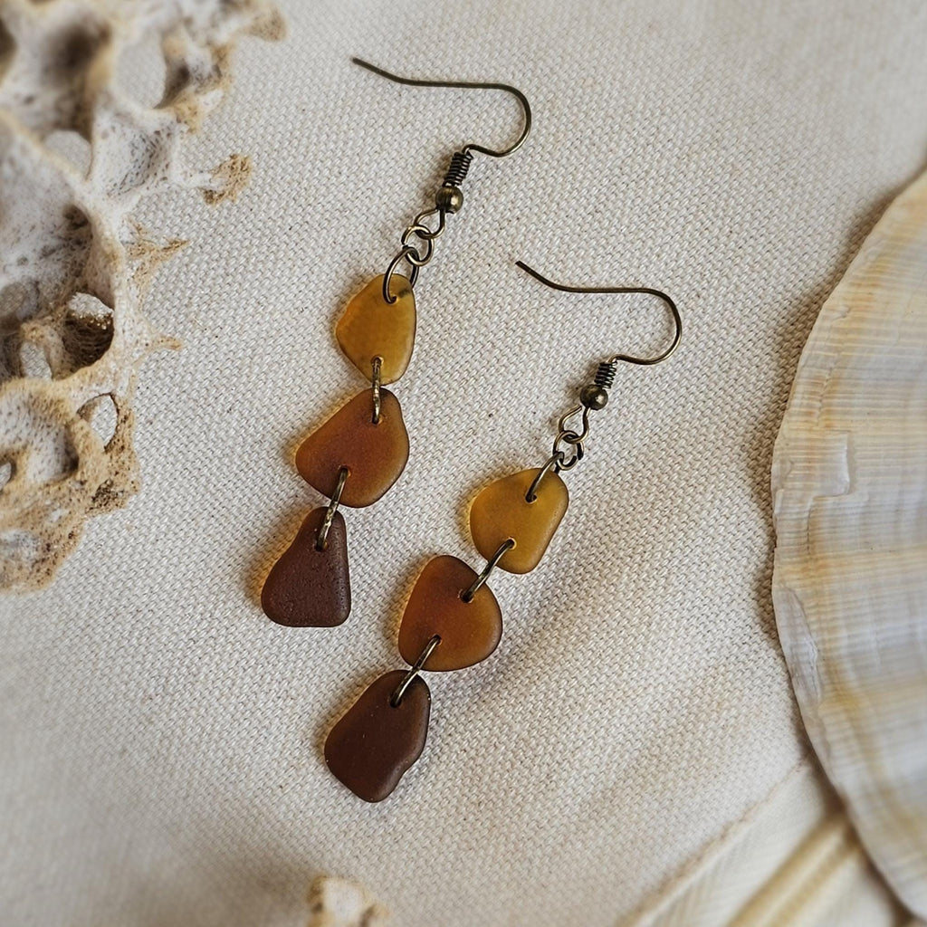 Pair of brown seaglass earrings on a textured beige surface with shells.