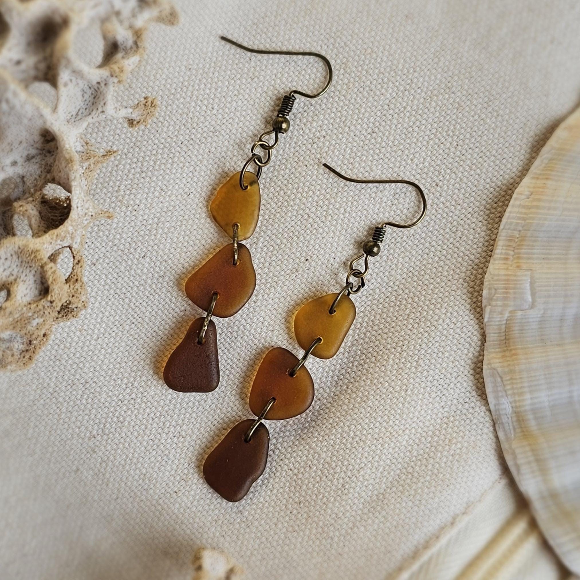 Pair of brown seaglass earrings on a textured beige surface with shells.